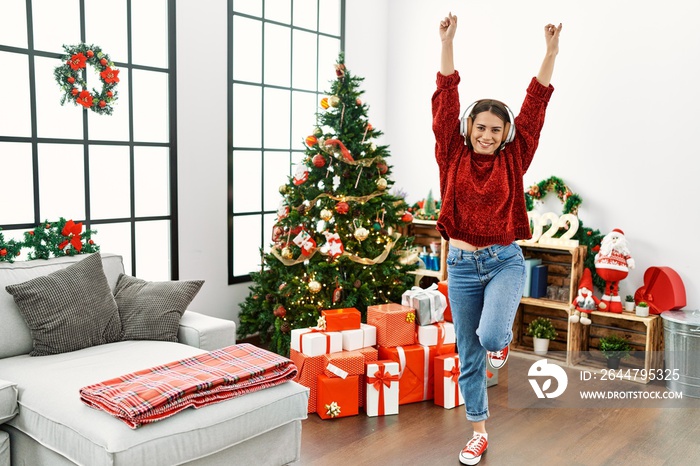 Young hispanic girl dancing using headphones standing by christmas tree at home.