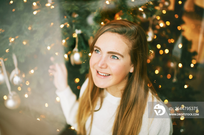 Young pretty girl looking up throught the window, christmas tree on background