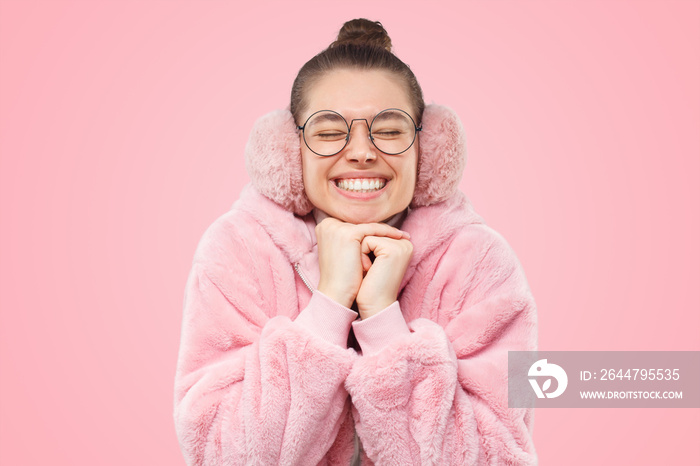 Girl in pink warm coat and earmuffs, holding hands together wishing dreams come true on Christmas