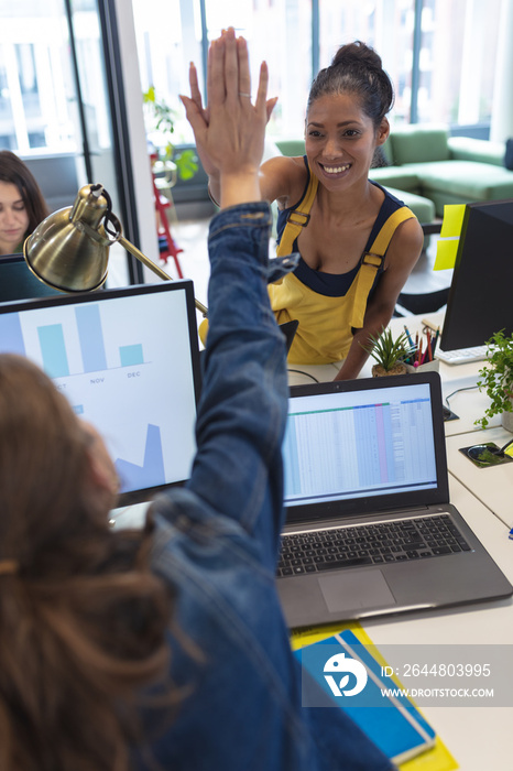 Two diverse female creative colleagues sitting at desk high-fiving and smiling