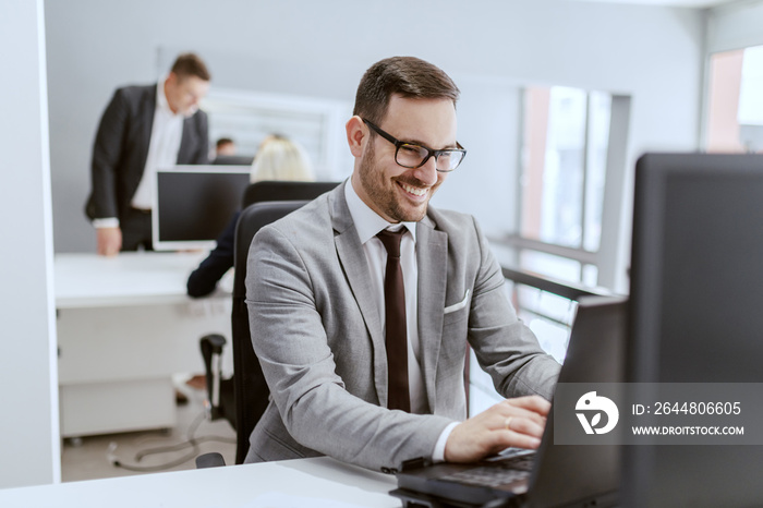 Smiling Caucasian businessman in suit and with eyeglasses sitting at his workplace and using compute