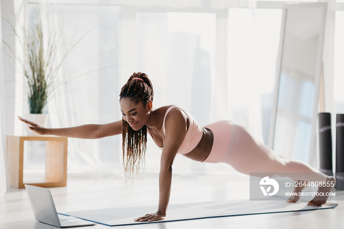 Fit young black woman standing in plank with outstretched arm, following online video tutorial on la