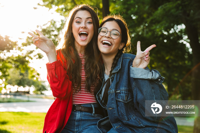 Image of two laughing girls gesturing peace sing and hugging while sitting on railing in green park