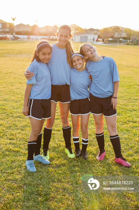 Portrait of smiling girls with arms around standing on soccer field against clear sky during sunset