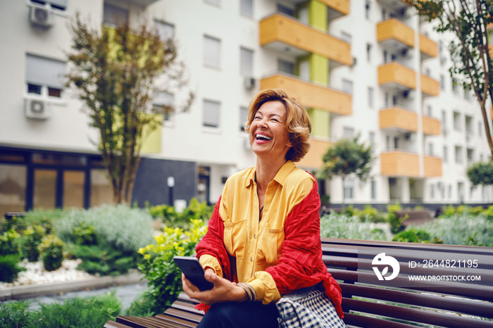 Gorgeous smiling caucasian fashionable senior woman with blonde short hair sitting on bench in park 