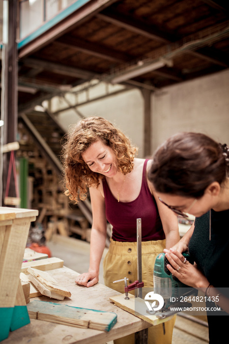 Smiling carpenter looking at female colleague cutting wood with jigsaw in workshop