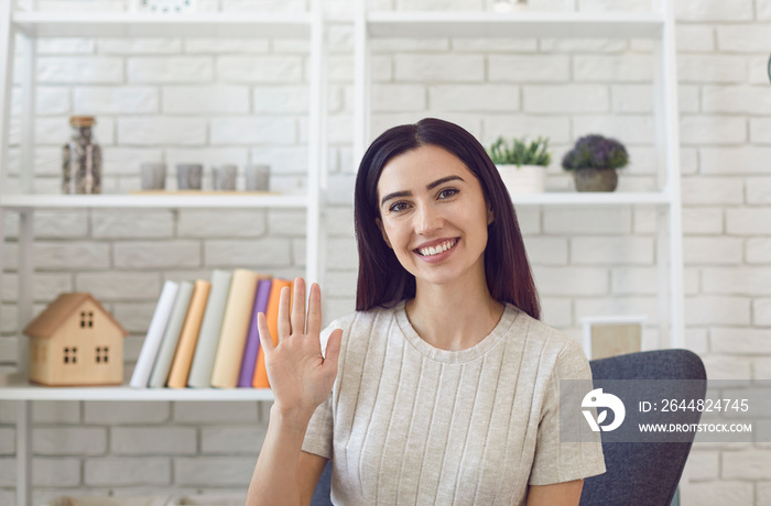 Smiling young woman in sitting on chair in modern office waving hand at camera