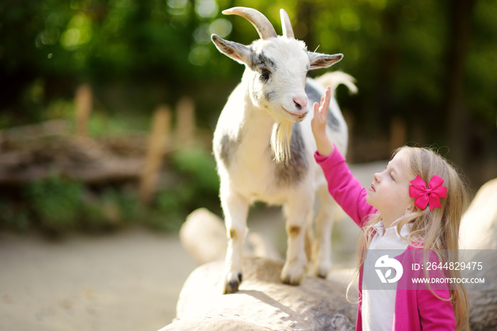 Cute little girl petting and feeding a goat at petting zoo. Child playing with a farm animal on sunn