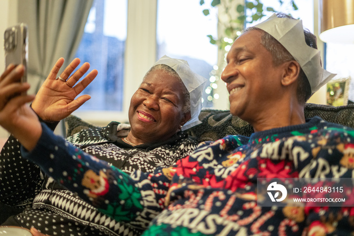 Senior couple sitting on sofa wearing Christmas decorations and waving to phone