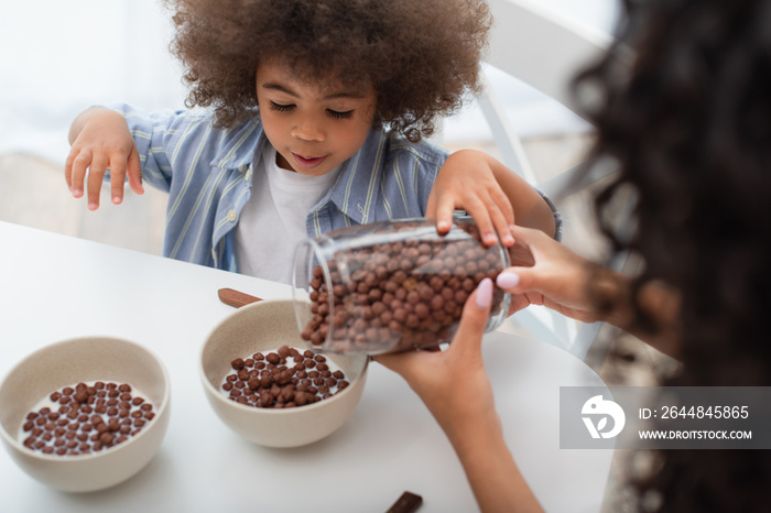 African american woman pouring cereal in bowls with milk near kid in kitchen.