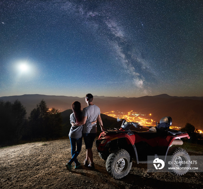 Rear view of happy young couple with atv quad motorbike on the top of mountain, boy and girl embraci