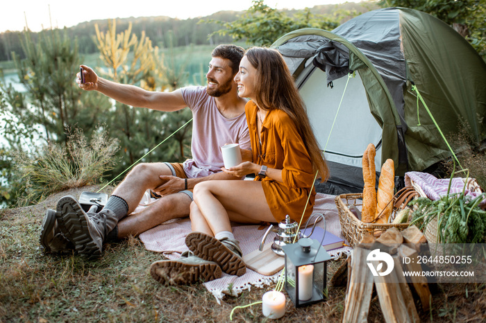 Young couple taking selfie photo, having fun during the picnic near the tent in the forest