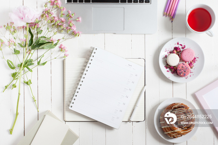 Feminine flat lay workspace with laptop, cup of tea, planner, macarons and flowers on white wooden t