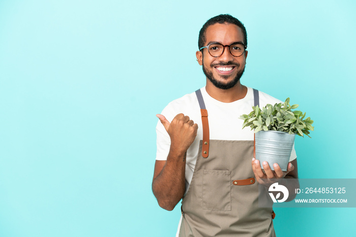 Gardener latin man holding a plant isolated on blue background pointing to the side to present a pro