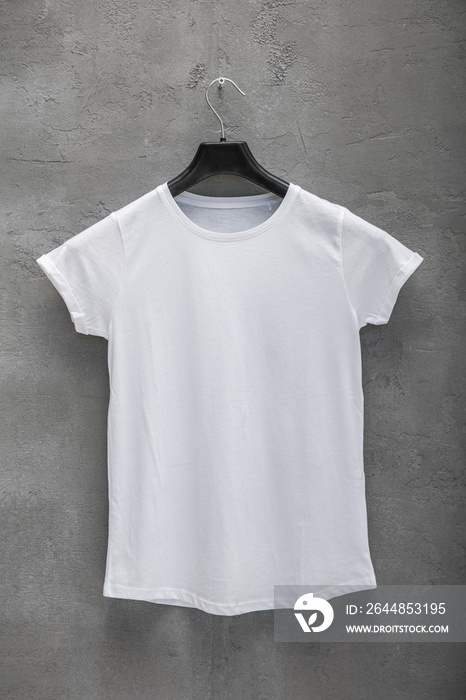 衣架上的女性白色棉质t恤正面，背景是混凝土墙。t恤