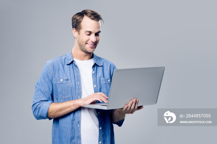 Portrait of happy smiling attractive man in blue smart casual wear, working with laptop, against gre