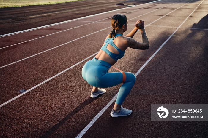 Attractive athlete girl in stylish sportswear squatting with rubber band during training on stadium