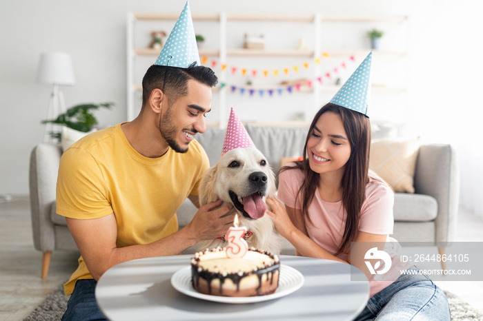 Joyful diverse spouses celebrating their dogs birthday with tasty cake, wearing festive hats at hom