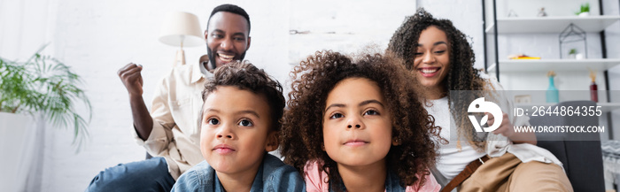 concentrated african american siblings watching tv near cheerful parents showing win gesture, banner