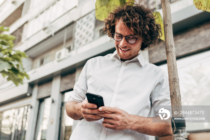 Outdoor shot of happy entrepreneur man in white shirt with transparent eyeglasses messaging on mobil