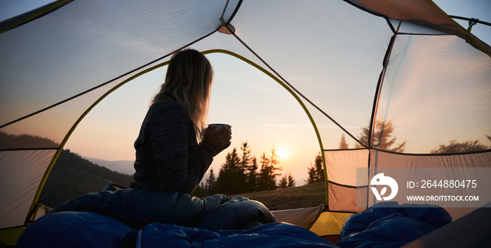 帐篷营地的早晨。孤独的女旅行者坐在帐篷里，手里拿着温暖的杯子的剪影
