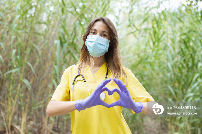 Mujer enfermera joven vestida con uniforme amarillo, guantes y mascarilla, forma un corazón con sus 