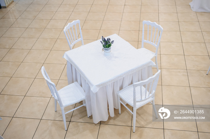 lateral view of a square table with white tablecloth and a little vase with flower on it surrounded by four white chairs in a banquet hall