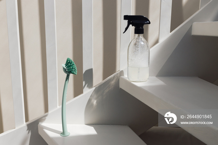 A green cleaning brush and a spray glass bottle with liquid standing on white staircase pointing at each other on a sunny day with shadows. Household utensil creative concept.