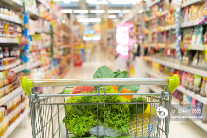 Fresh fruits and vegetables in shopping cart or trolly with aisle and label products on shelves at supermarket or grocery store in mall. Food and lifestyle