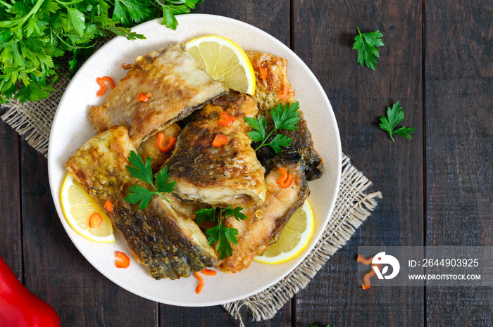 Pieces of roasted carp with lemon and greens on a plate on a dark wooden background. Top view.