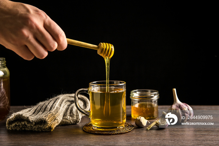 Man hand holding wooden honey dipper, honey spoon on top of glass of tea/ medicine and dripping honey in hot tea. Knitted socks, small jar of honey, garlic on wooden table against black background.