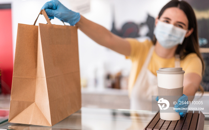 Young woman wearing face mask while serving takeaway breakfast and coffee inside cafeteria restaurant - Worker preparing delivery food inside bakery bar during coronavirus period - Focus on right hand