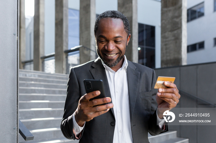 Happy and successful african american man in business suit outside office building holding bank credit card and phone, businessman shopping online and transferring money using app.