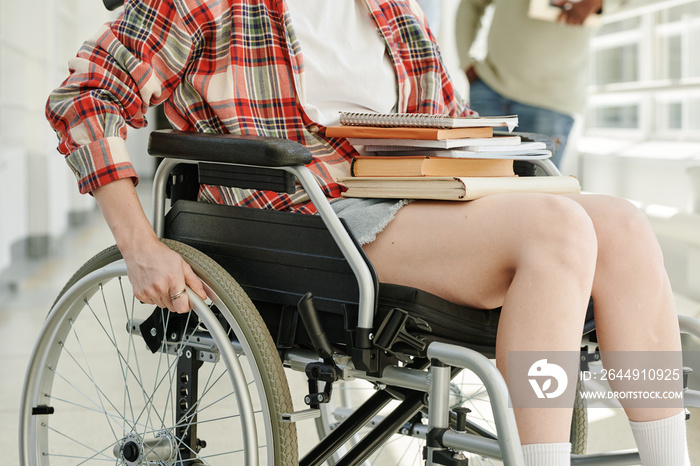 Close-up of teenage girl with stack of books and copybooks on her knees rotating wheels of wheelchair while moving along college corridor