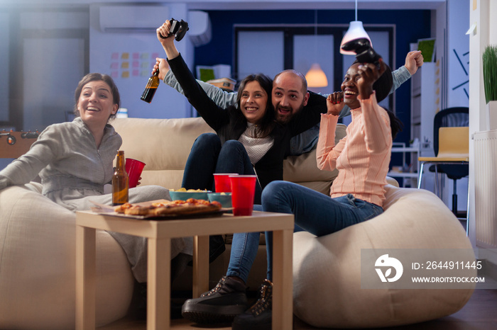 Multi ethnic friends celebrate together after work at office while playing game on tv console with controller joysticks. Diverse group of colleagues having fun gaming at party celebration