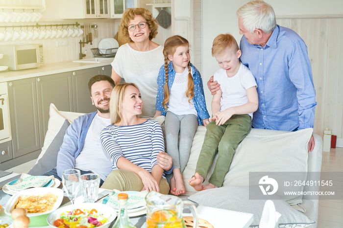 Portrait of happy two generation family enjoying dinner together in modern apartment posing on couch with two adorable children in sunlight