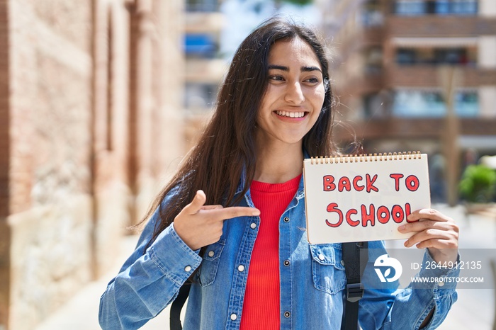 Young hispanic girl student pointing with finger to back to school notebook at street