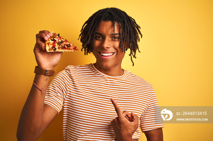 Afro american man with dreadlocks eating slice of pizza over isolated yellow background very happy pointing with hand and finger