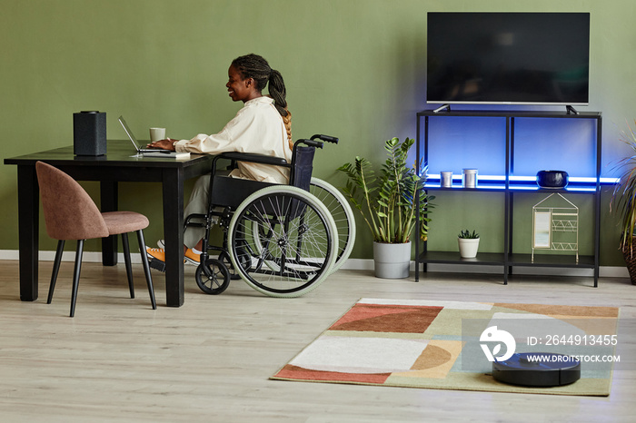 Side view portrait of young African American woman using wheelchair while working at home office with smart home accessible devices