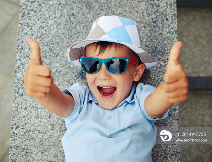 Small boy in sunglasses giving thumbs up while lying on street s