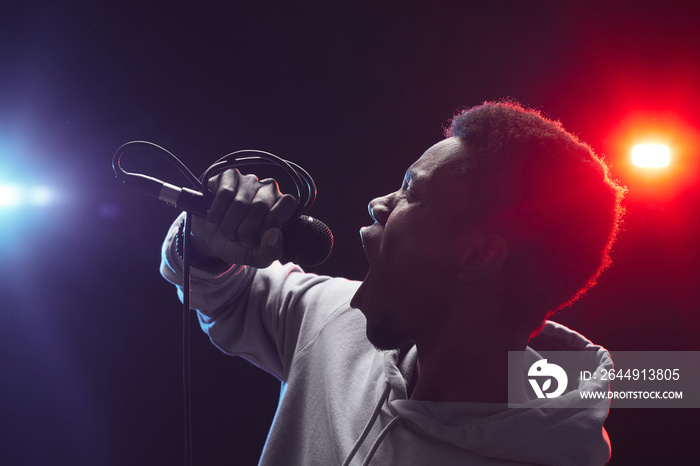 Side view portrait of young African-American man singing to microphone emotionally while standing on stage in lights