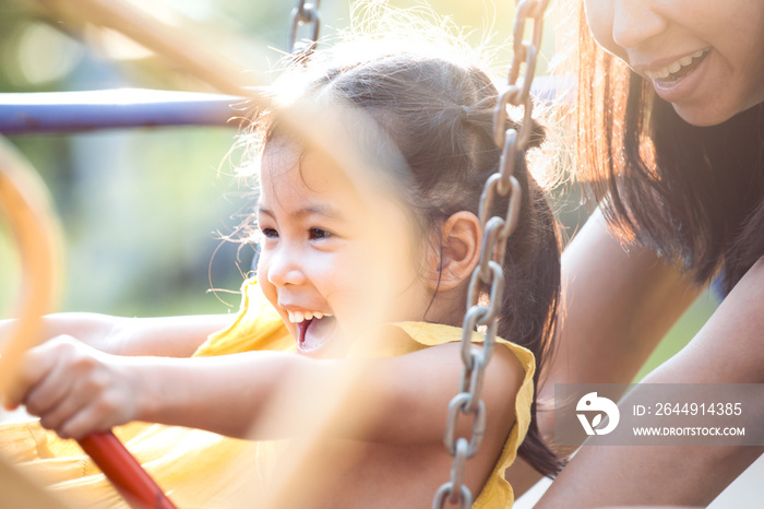 Happy asian little child girl having fun to ride on swings when mother pushing her swings in playground in the sunset time