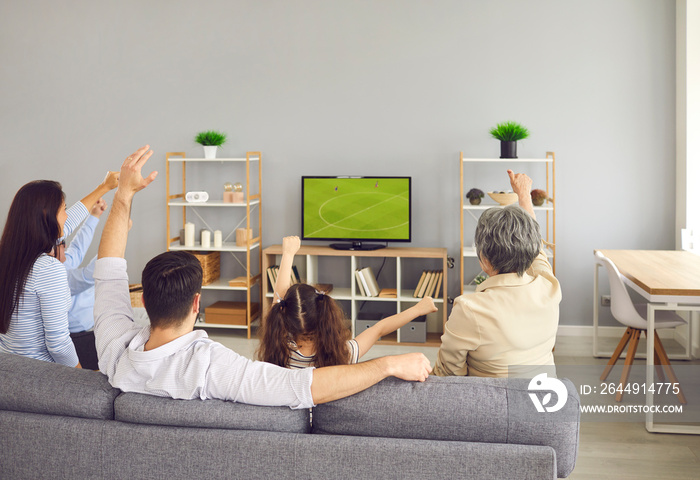 Super excited and happy big family team watch football match together on the couch at home. Rear view of young and older family members sitting with their arms raised while watching a match on TV.