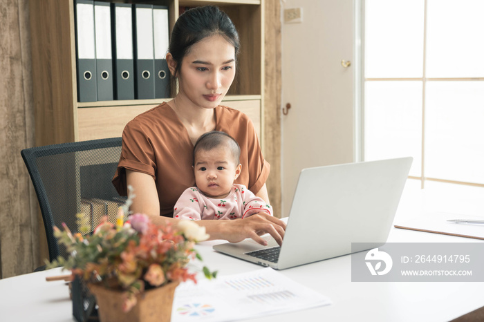 Asia mother working online with a laptop holding her baby daughter at home office. Serious single mom with child. Asian woman working from home, while in quarantine isolation during Covid-19 crisis.
