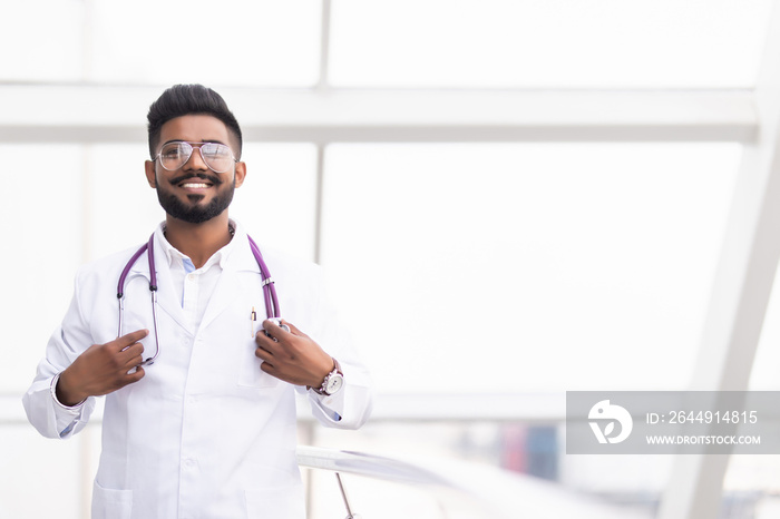 Portrait of a smiling indian male medical doctor in uniform, hospital building at background.