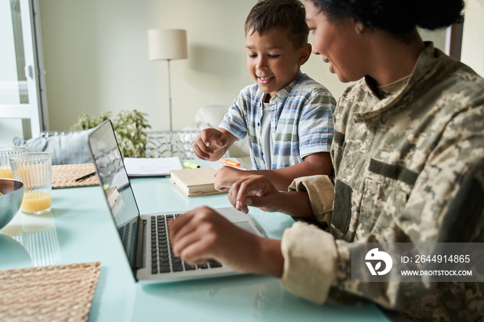 Boy looking at the laptop while his soldier mother in military uniform working