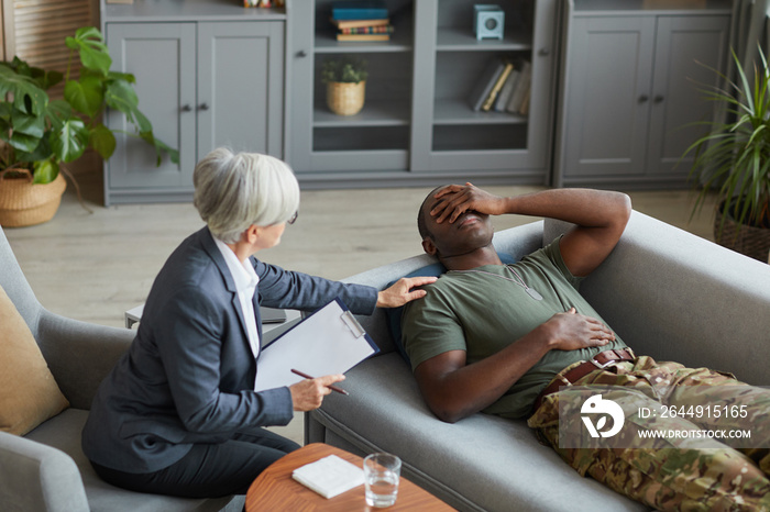 African military man lying on sofa in depression while psychologist supporting him during therapy at office