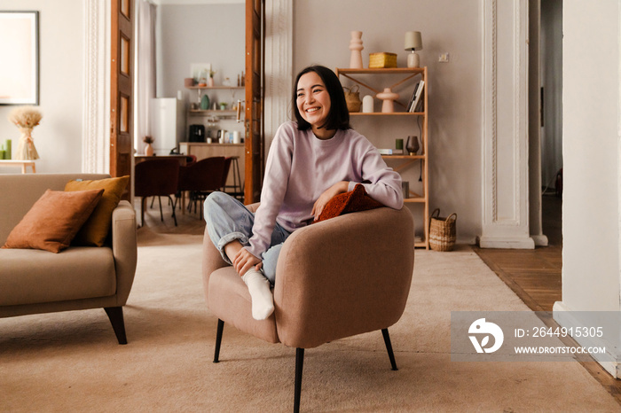 Beautiful asian woman laughing while sitting on armchair in living room