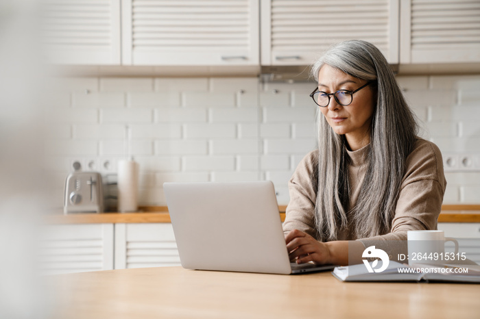 Mature grey woman working with laptop while sitting in kitchen at home