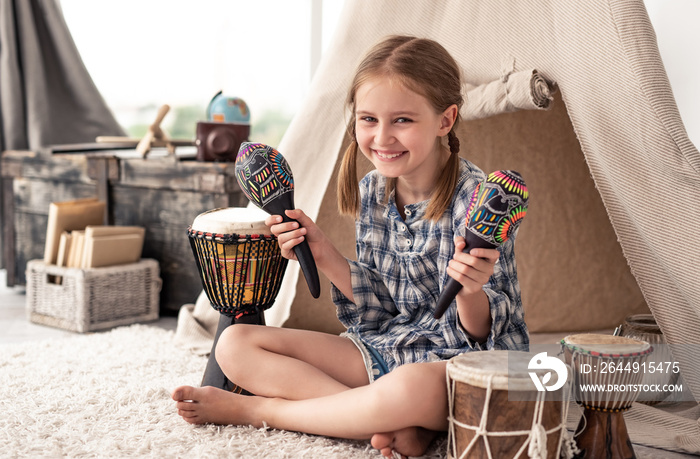 Smiling little girl shaking music maracas sitting in room with djembe drums and wigwam